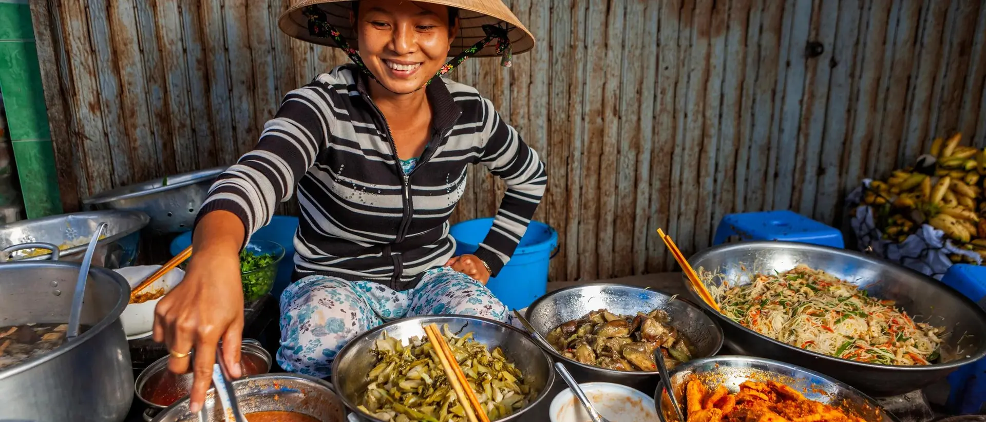 A smiling woman in a conical hat serves dishes from a variety of bowls at a vibrant market stall.