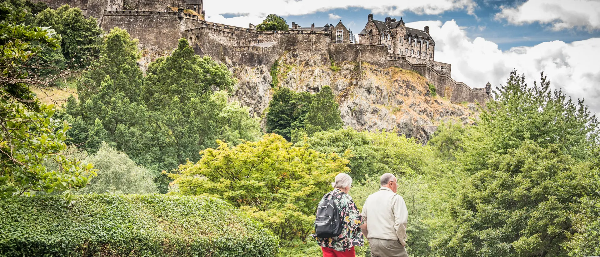 Two people stroll along a path, with Edinburgh Castle perched on a rocky hill in the background, surrounded by greenery.