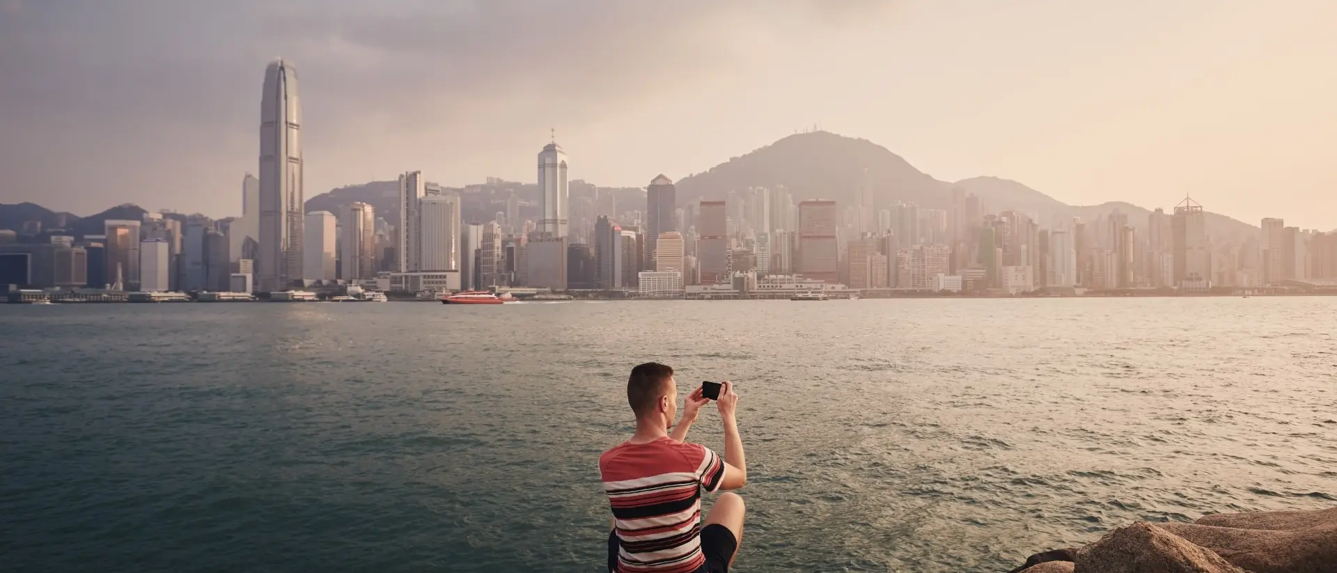 A person sitting on rocks by the water, taking a photo of the Hong Kong skyline at sunset.