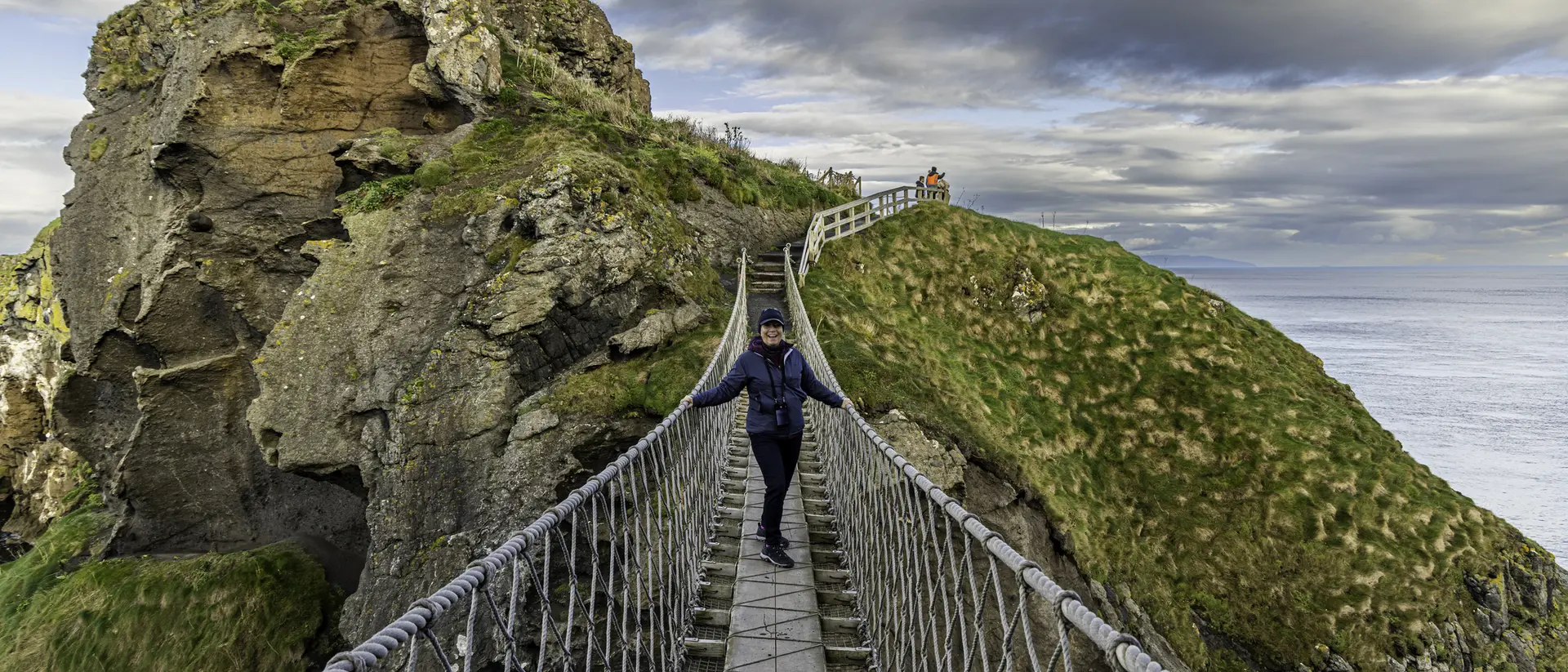 Person walking on a suspension bridge between cliffs, with grassy hills and a cloudy sky in the background.