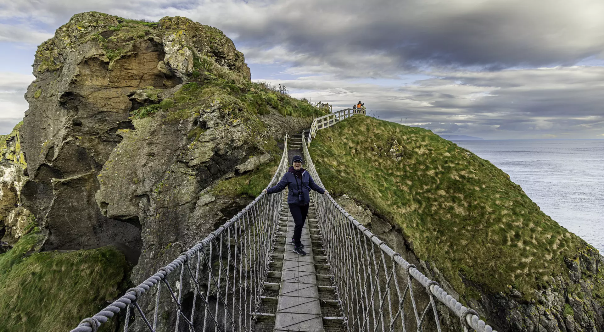 Person walking on a suspension bridge between cliffs, with grassy hills and a cloudy sky in the background.