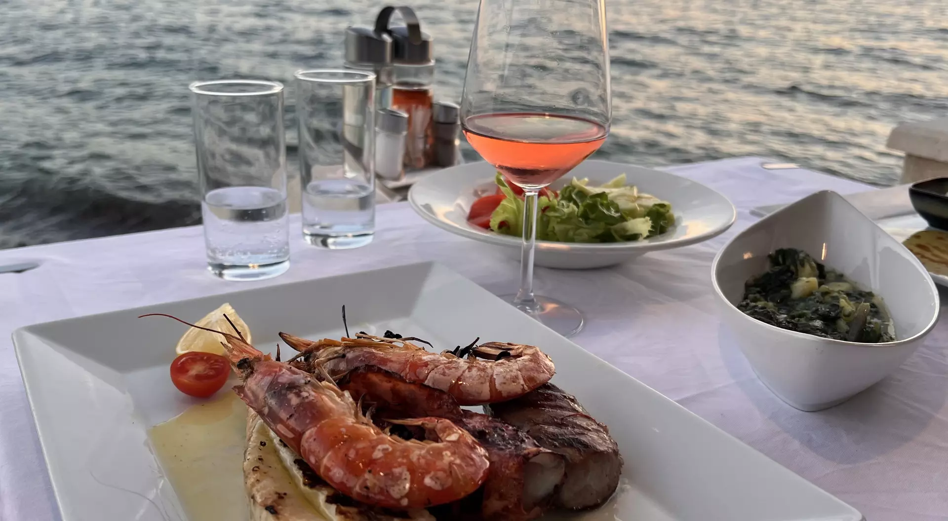 A plated seafood dish with shrimp and fish, a glass of rose wine, and salads, set on a table by the water at sunset.