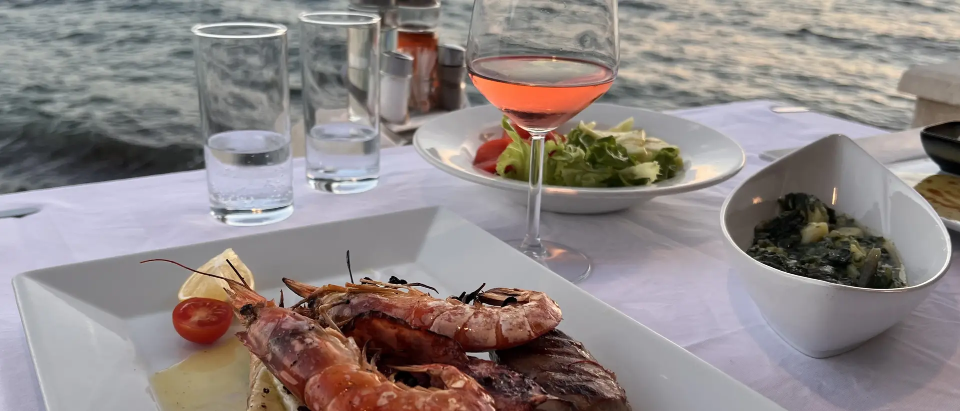 A plated seafood dish with shrimp and fish, a glass of rose wine, and salads, set on a table by the water at sunset.