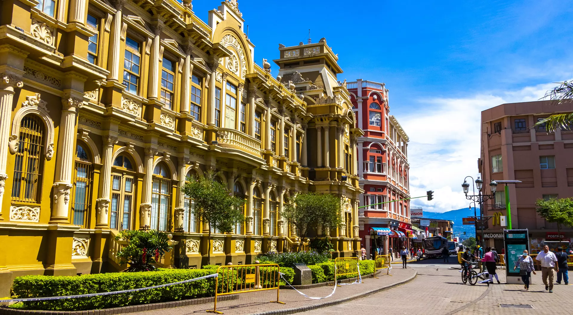 Historic buildings line a sunny street, with greenery and pedestrians enjoying the vibrant urban scene.