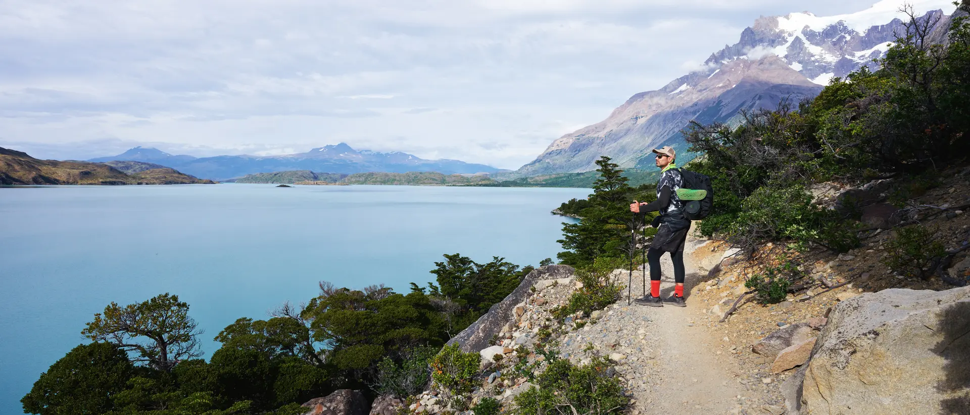 Hiker stands on a trail overlooking a turquoise lake with mountains in the background.