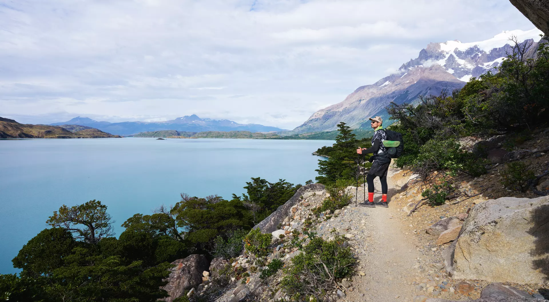 Hiker stands on a trail overlooking a turquoise lake with mountains in the background.