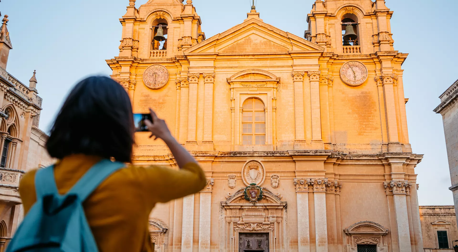 A person with a backpack takes a photo of a historic church bathed in golden light at sunset.