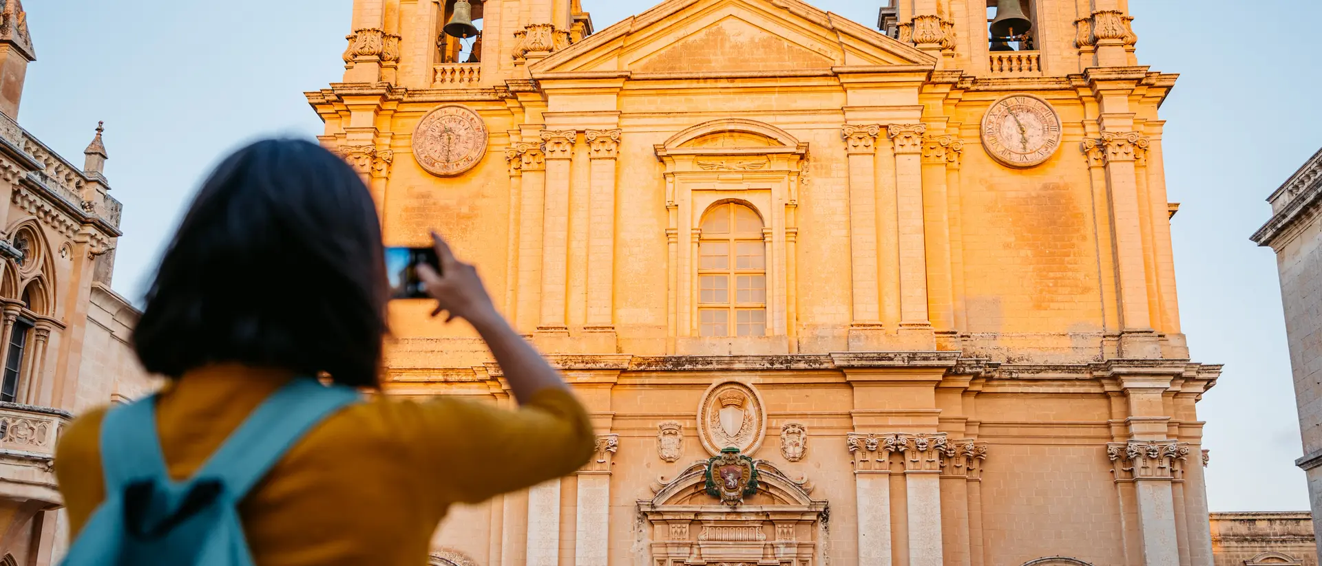 A person with a backpack takes a photo of a historic church bathed in golden light at sunset.