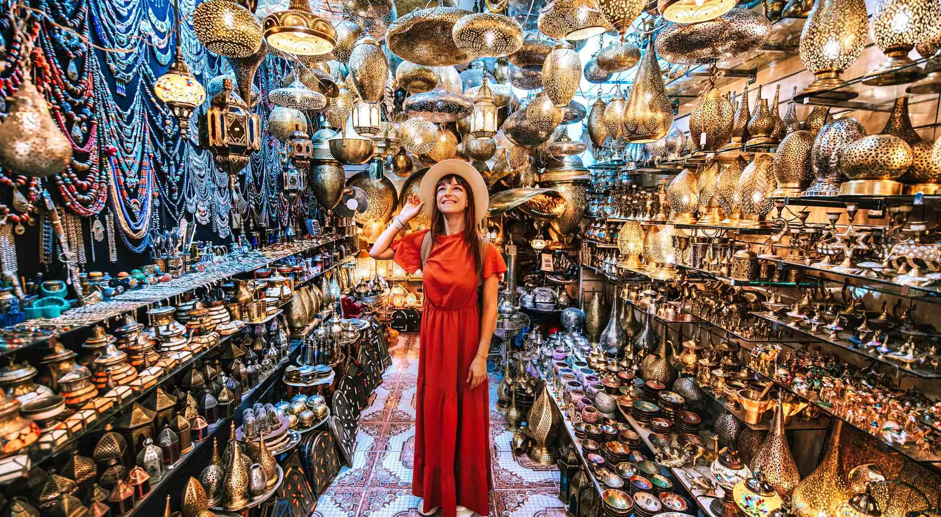 Person in a red dress and hat stands in a vibrant shop filled with intricate metal lanterns and decorative items.