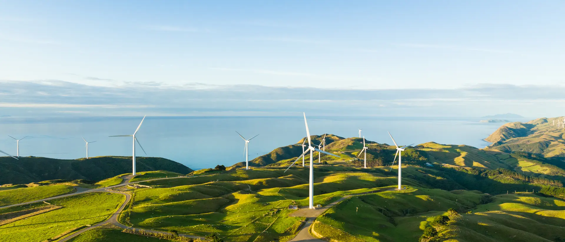 Aerial view of green hills with wind turbines overlooking the ocean and clear blue sky.