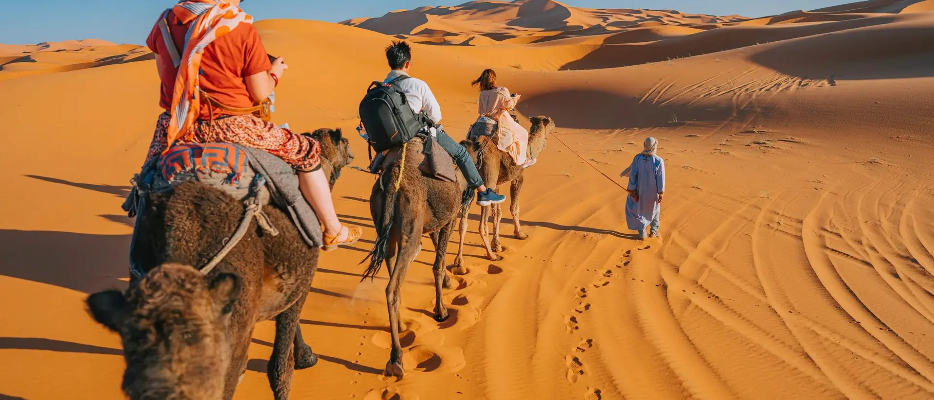 A group of people riding camels through golden sand dunes under a clear blue sky.