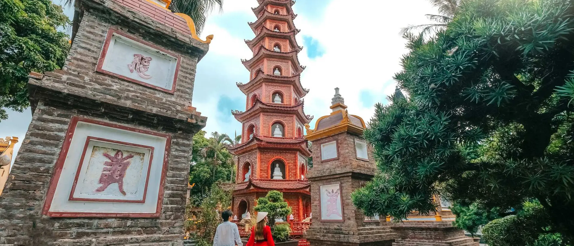 Couple walking towards a tall pagoda surrounded by greenery and traditional stone structures.
