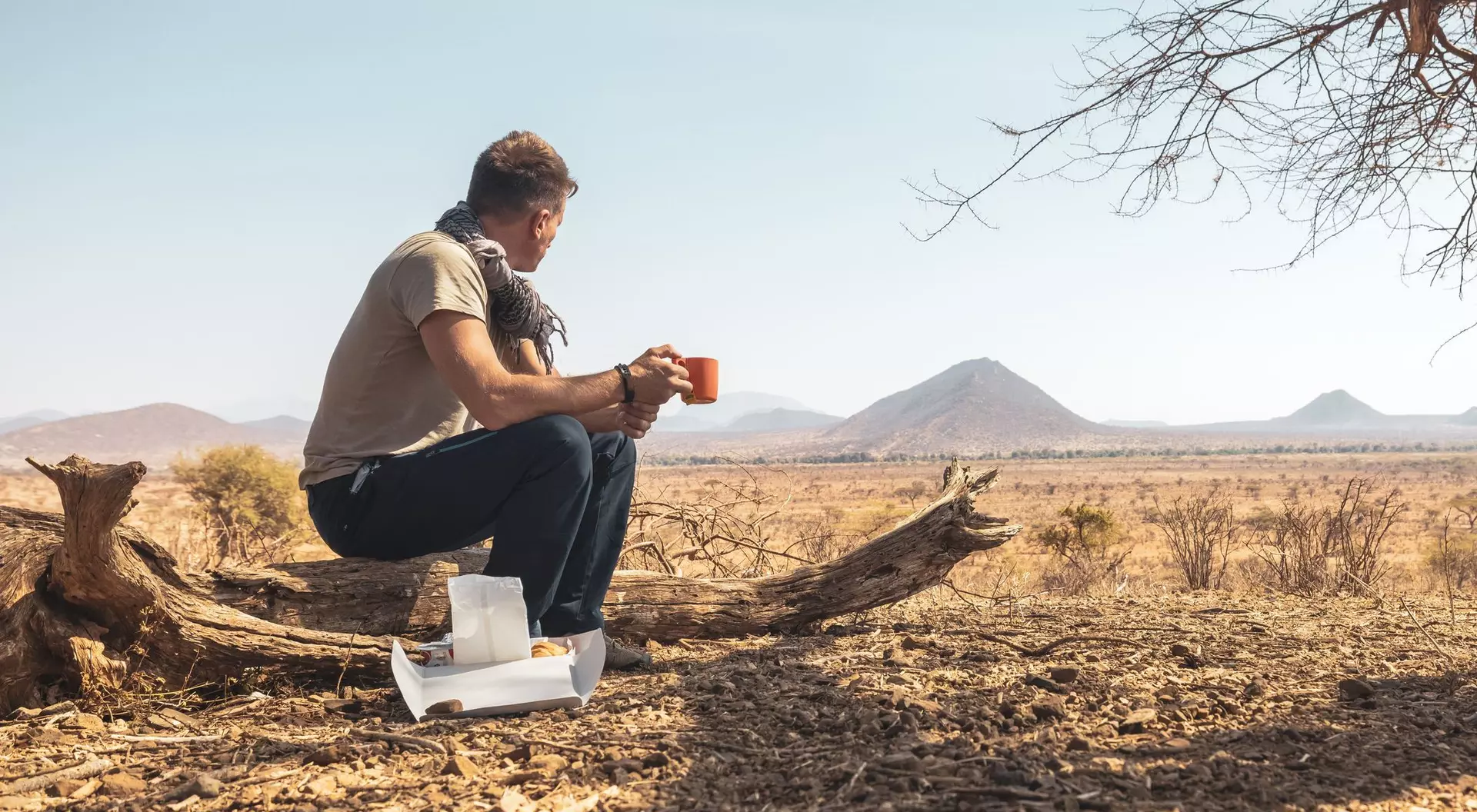 A person sits on a log in a vast, arid landscape, holding a cup and looking out at distant mountains.