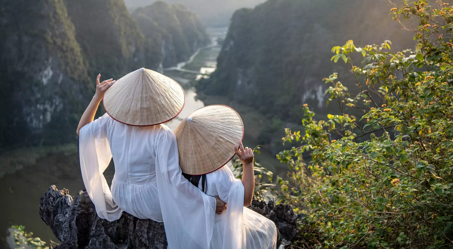 Two women in traditional conical hats sit on a rocky ledge, overlooking a scenic river and mountains at sunset.