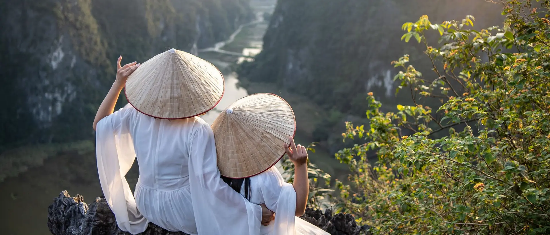 Two women in traditional conical hats sit on a rocky ledge, overlooking a scenic river and mountains at sunset.