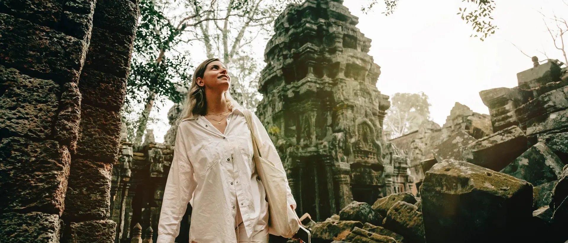 A woman in white clothing explores ancient ruins surrounded by overgrown trees and stone structures.