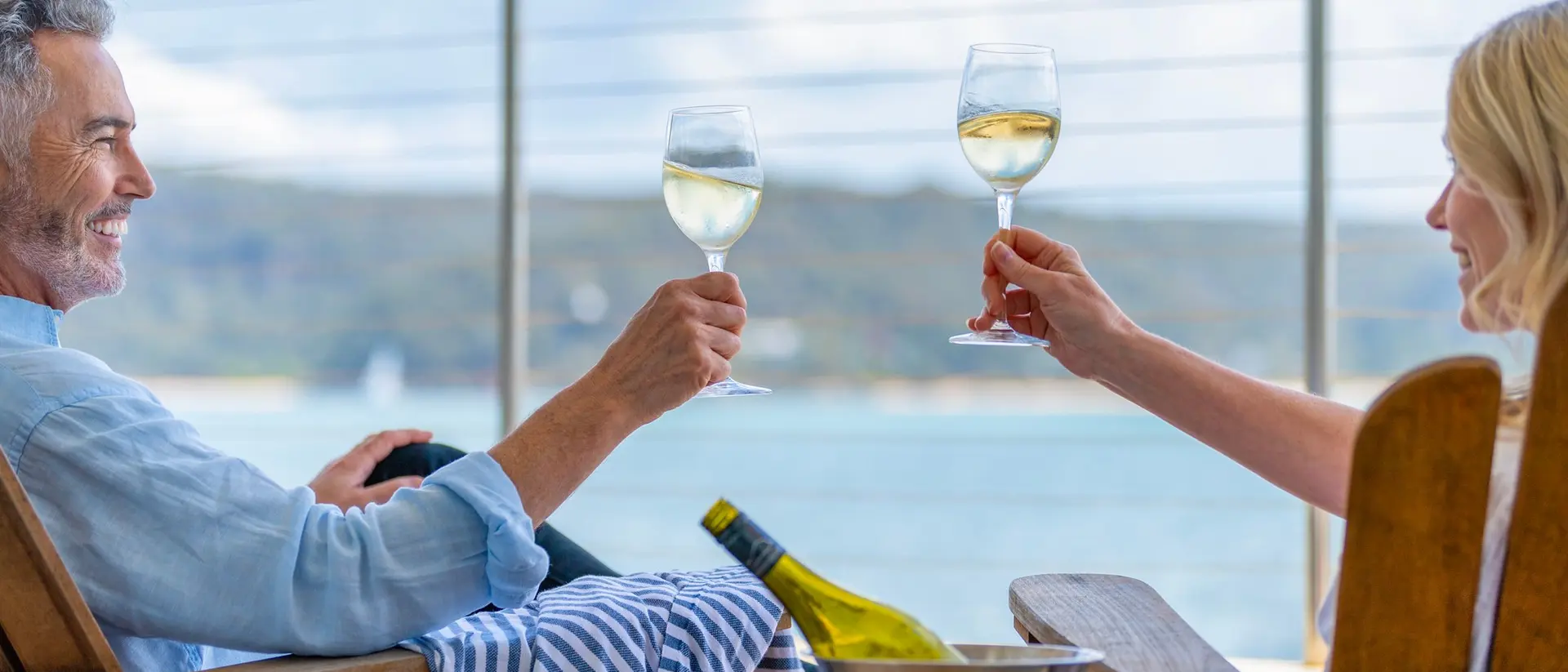 A couple toasting with wine glasses while sitting on a balcony overlooking a scenic water view.