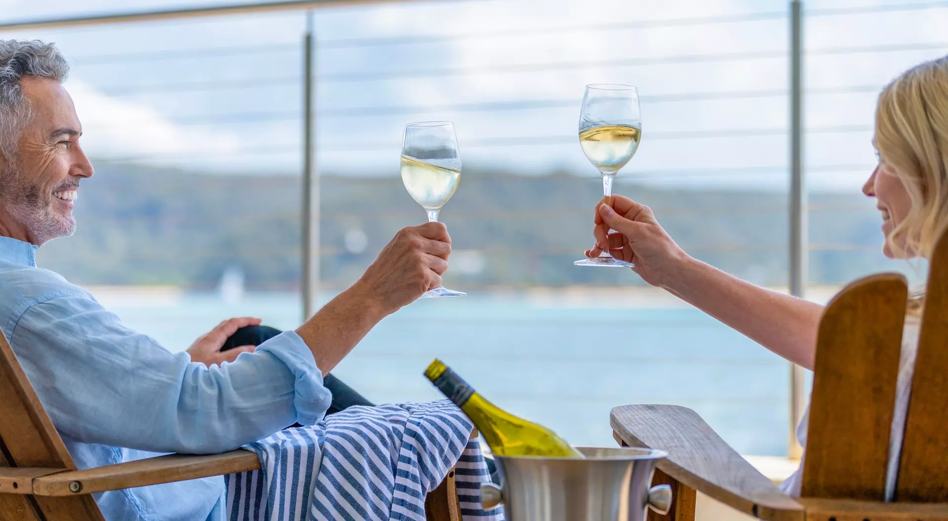 A couple toasting with wine glasses while sitting on a balcony overlooking a scenic water view.