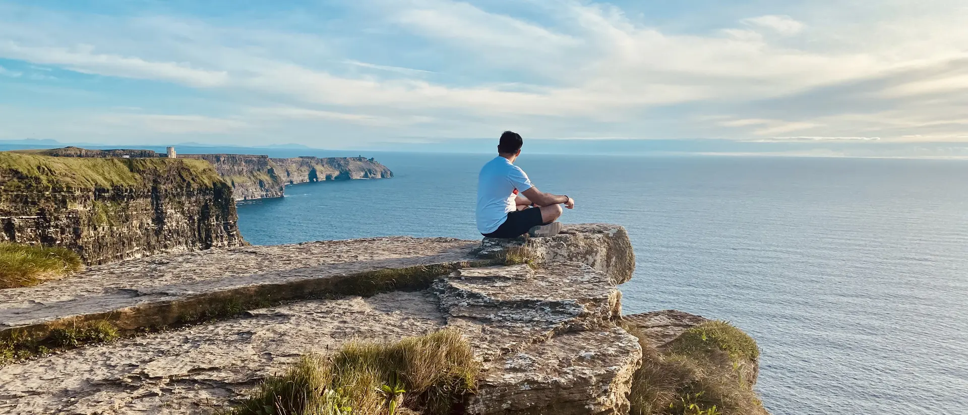 A person sits on a rocky cliff, overlooking a calm sea under a clear sky.