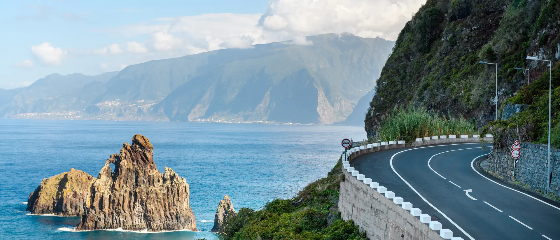 A coastal road curves along the cliffs, with rocky outcrops and mountains visible across the blue ocean.