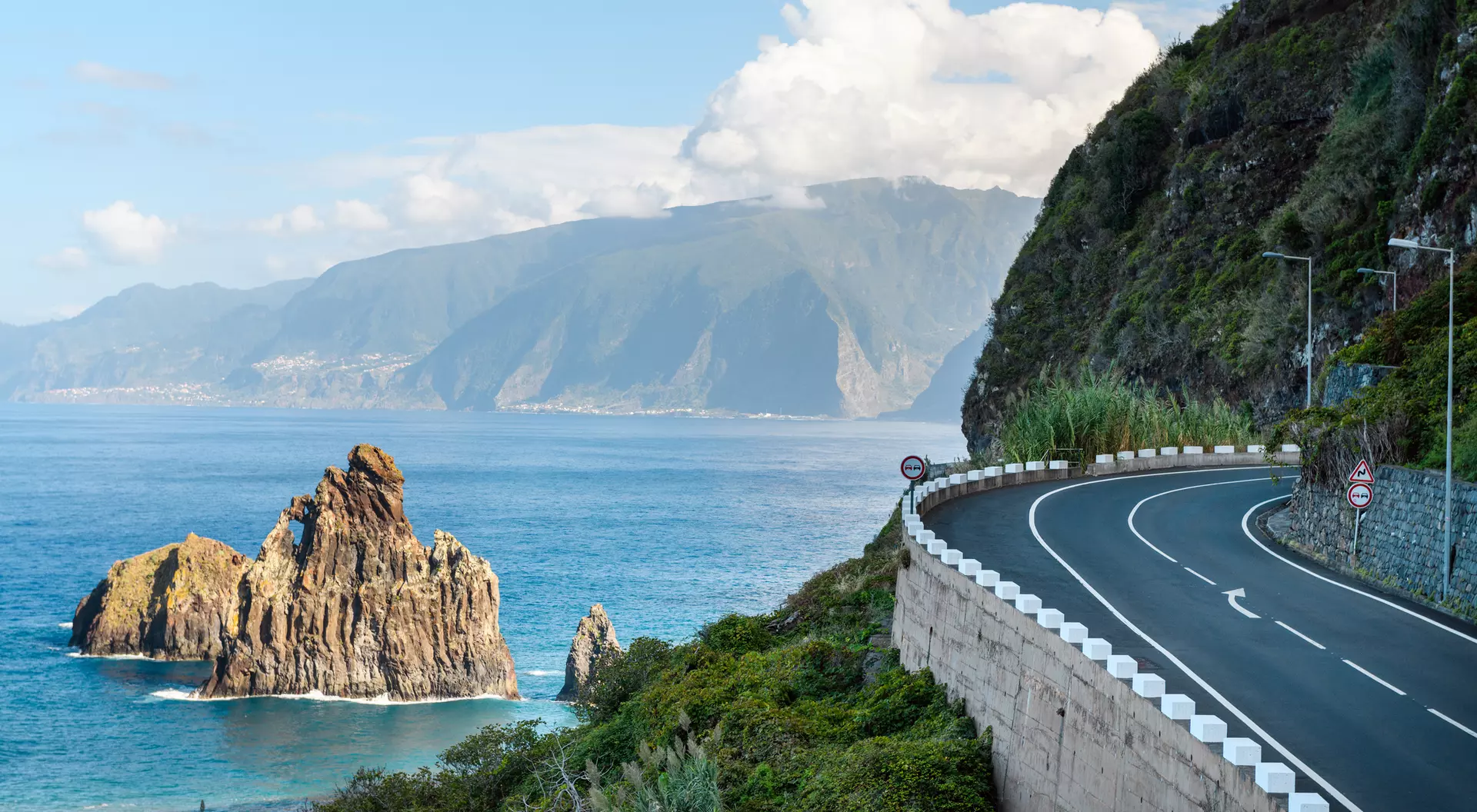 A coastal road curves along the cliffs, with rocky outcrops and mountains visible across the blue ocean.