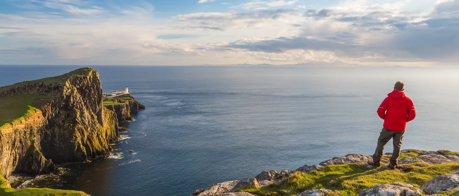 A person in a red jacket stands on rocky cliffs, gazing at the ocean and a distant lighthouse under a clear sky.