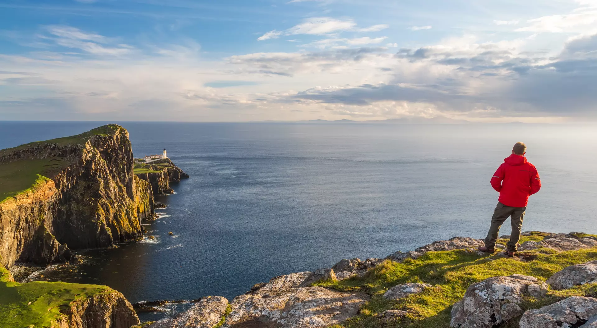 A person in a red jacket stands on rocky cliffs, gazing at the ocean and a distant lighthouse under a clear sky.
