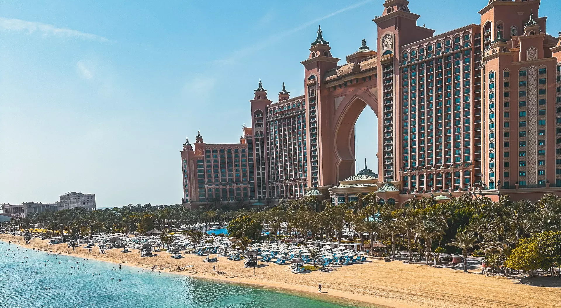 Luxury beachfront resort with palm trees, blue water, and a tall, ornate hotel under a clear blue sky.