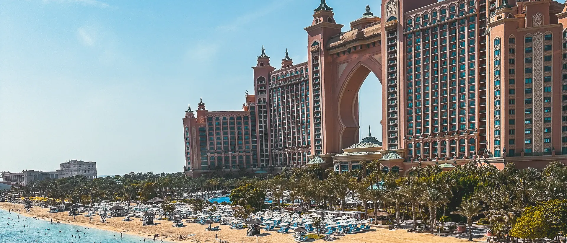 Luxury beachfront resort with palm trees, blue water, and a tall, ornate hotel under a clear blue sky.