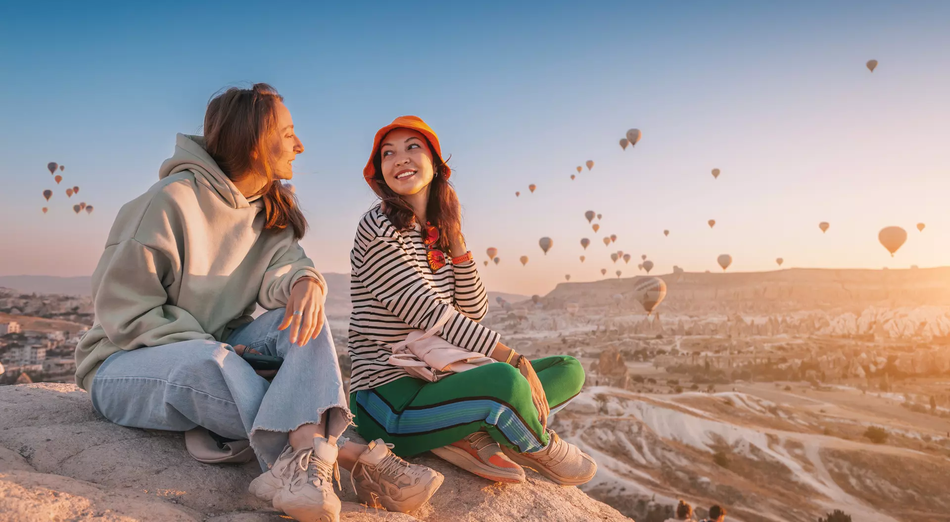 Two women sit on a rock, smiling and enjoying the view of colorful hot air balloons in the sky during sunset.
