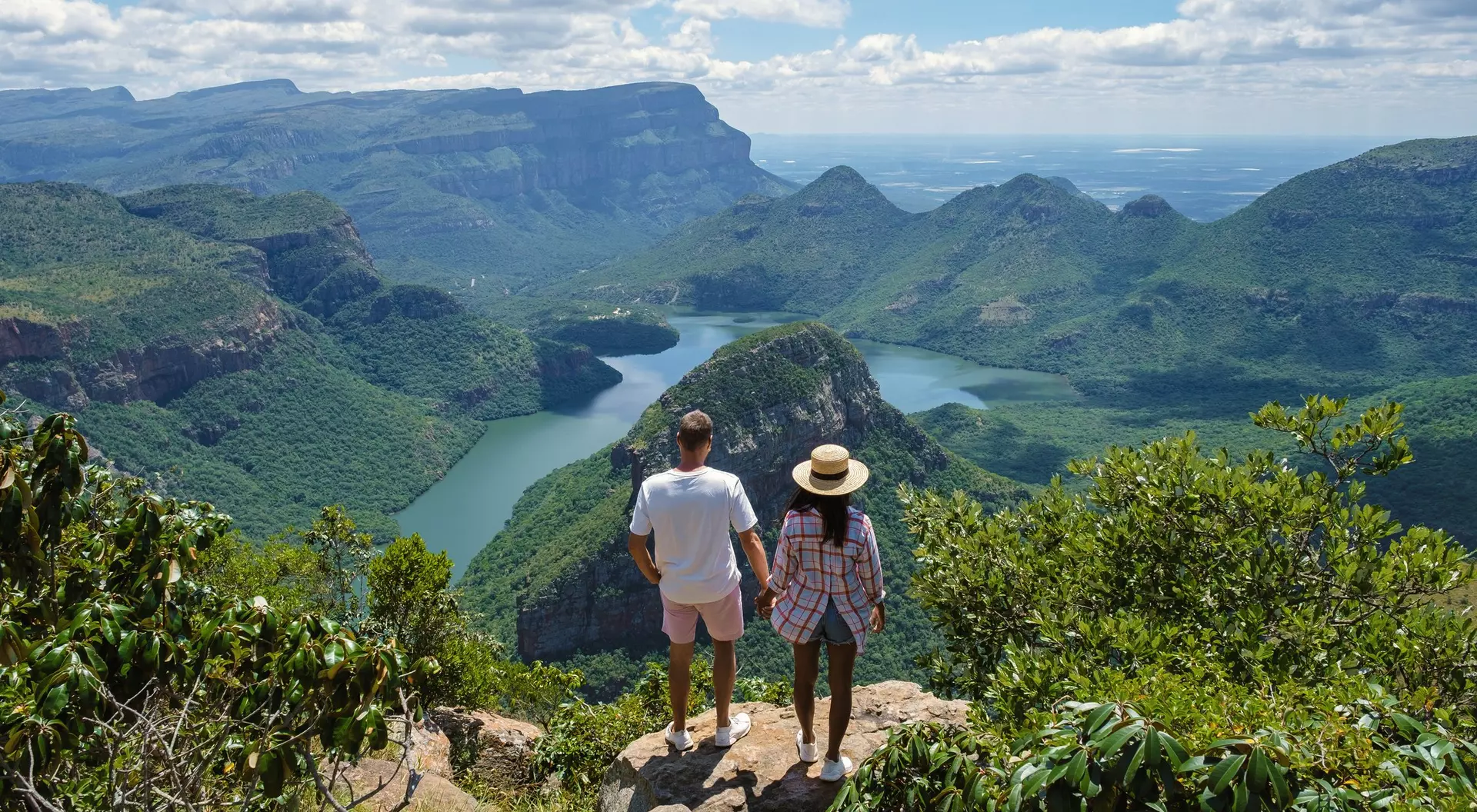 A couple stands hand-in-hand on a cliff, overlooking a lush green valley and a winding river under a bright blue sky.