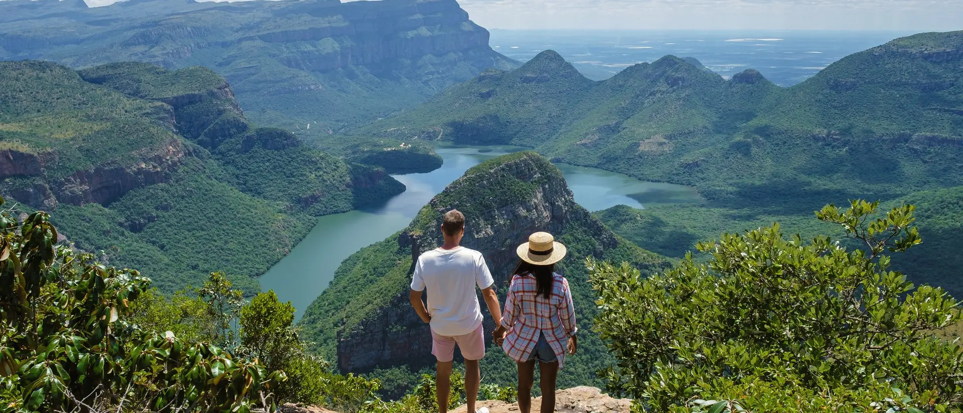 A couple stands hand-in-hand on a cliff, overlooking a lush green valley and a winding river under a bright blue sky.