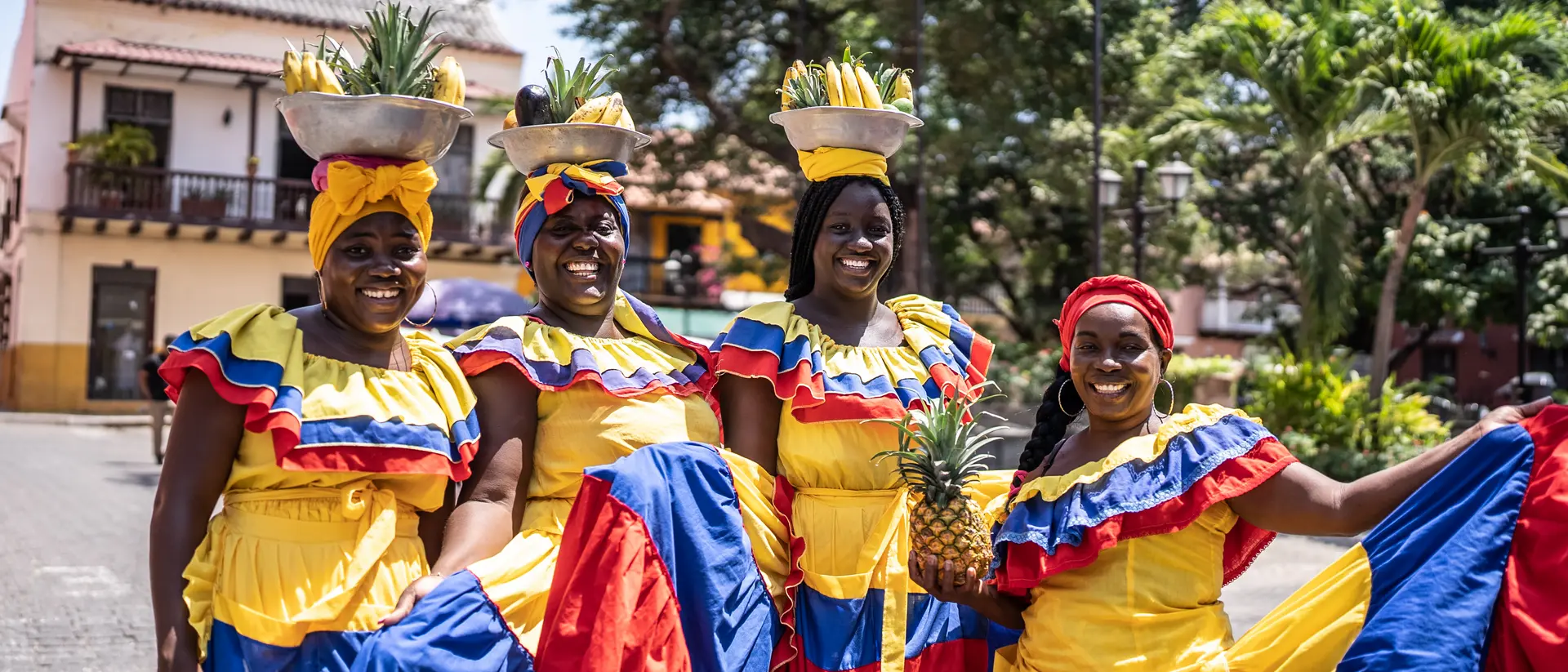 Four women in bright traditional dresses, each carrying a pineapple on their heads, pose happily outdoors.