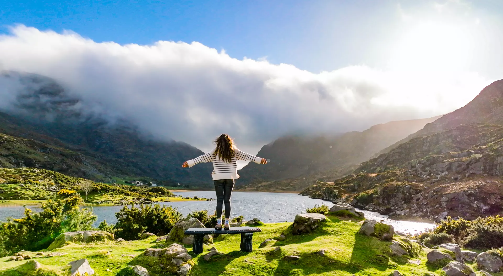 Person standing on a rock by a lake, arms outstretched, surrounded by mountains and a bright sky with clouds.