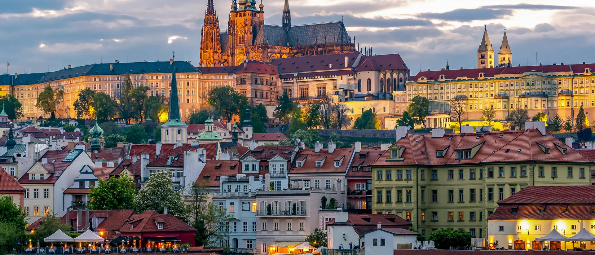 View of Prague Castle illuminated at sunset, overlooking colorful buildings along the Vltava River.