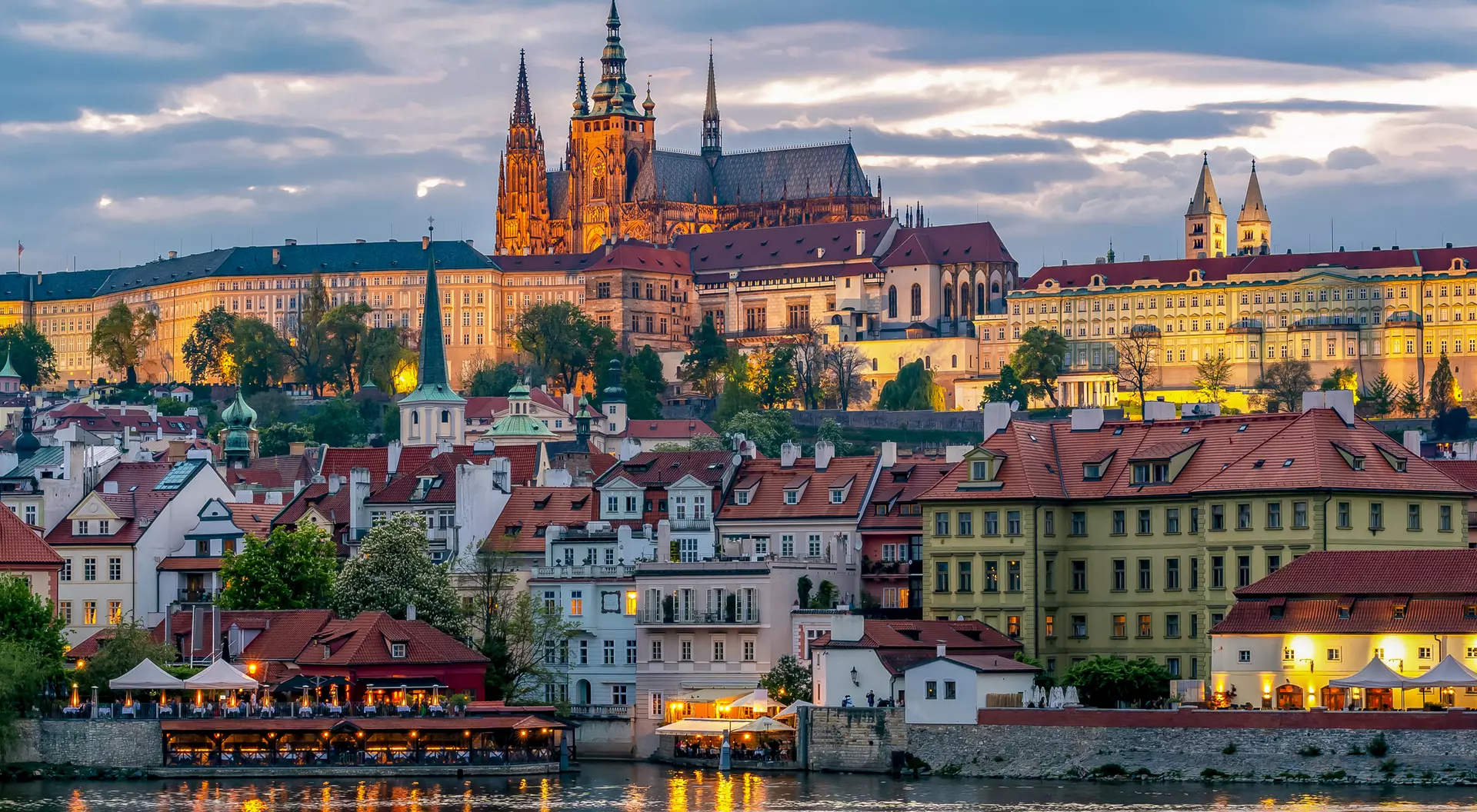 View of Prague Castle illuminated at sunset, overlooking colorful buildings along the Vltava River.