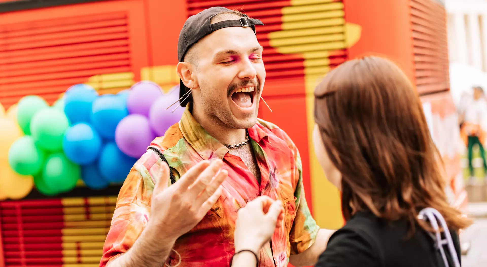 A person with colorful clothing and makeup joyfully interacts with another person against a vibrant, festive backdrop.