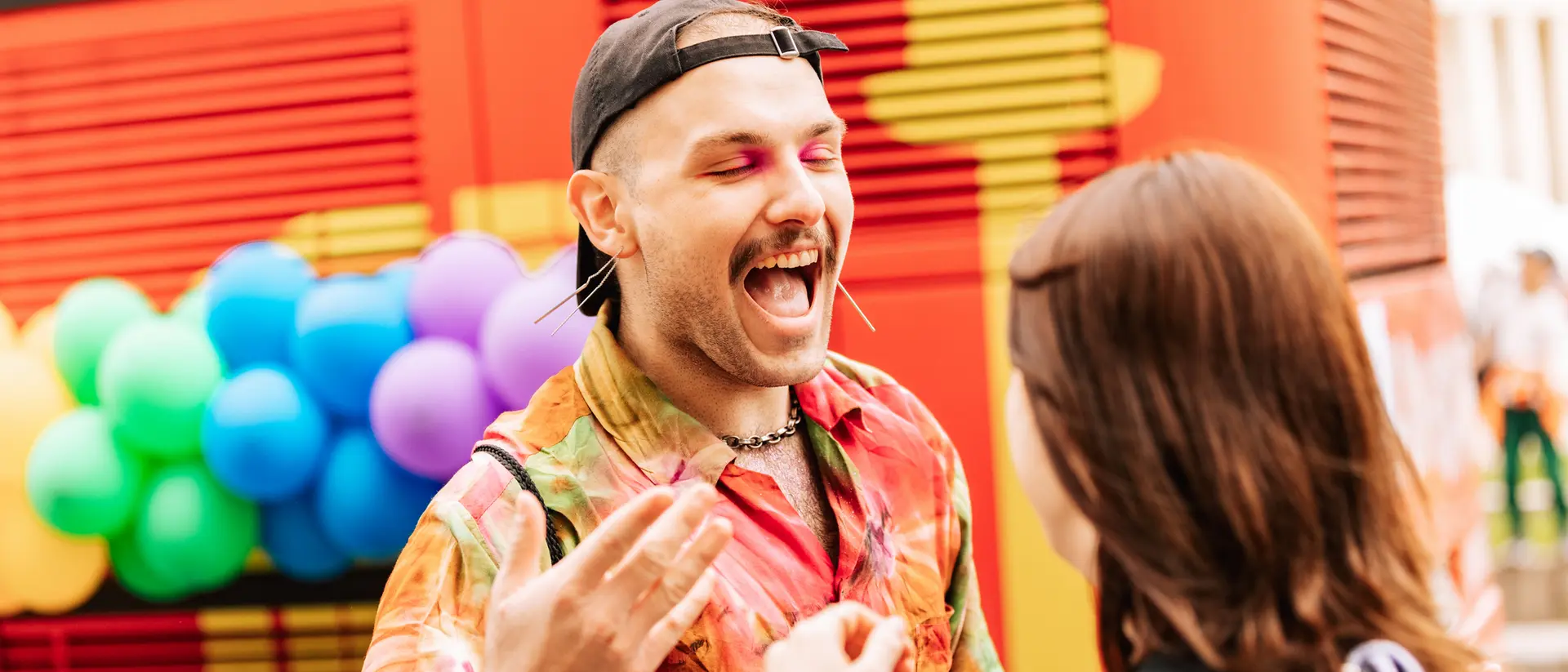 A person with colorful clothing and makeup joyfully interacts with another person against a vibrant, festive backdrop.