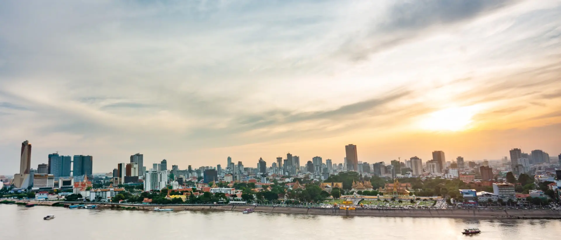 City skyline at sunset over a river, with clouds and boats visible on the water.