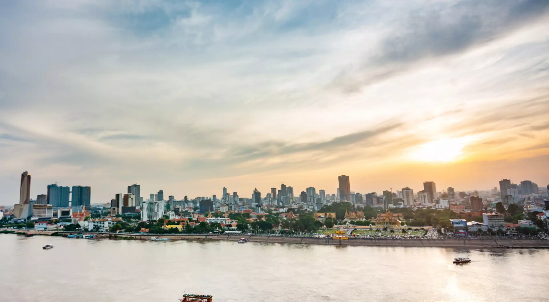City skyline at sunset over a river, with clouds and boats visible on the water.
