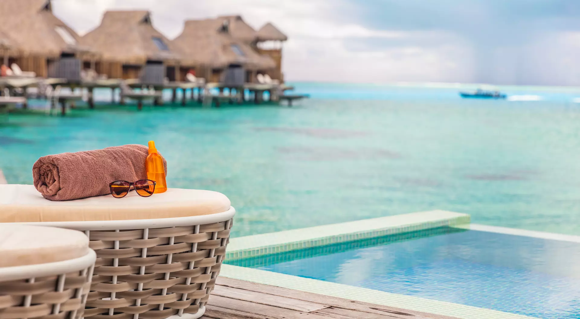 A poolside view featuring a rolled towel and sunglasses beside a turquoise sea and overwater bungalows in the background.