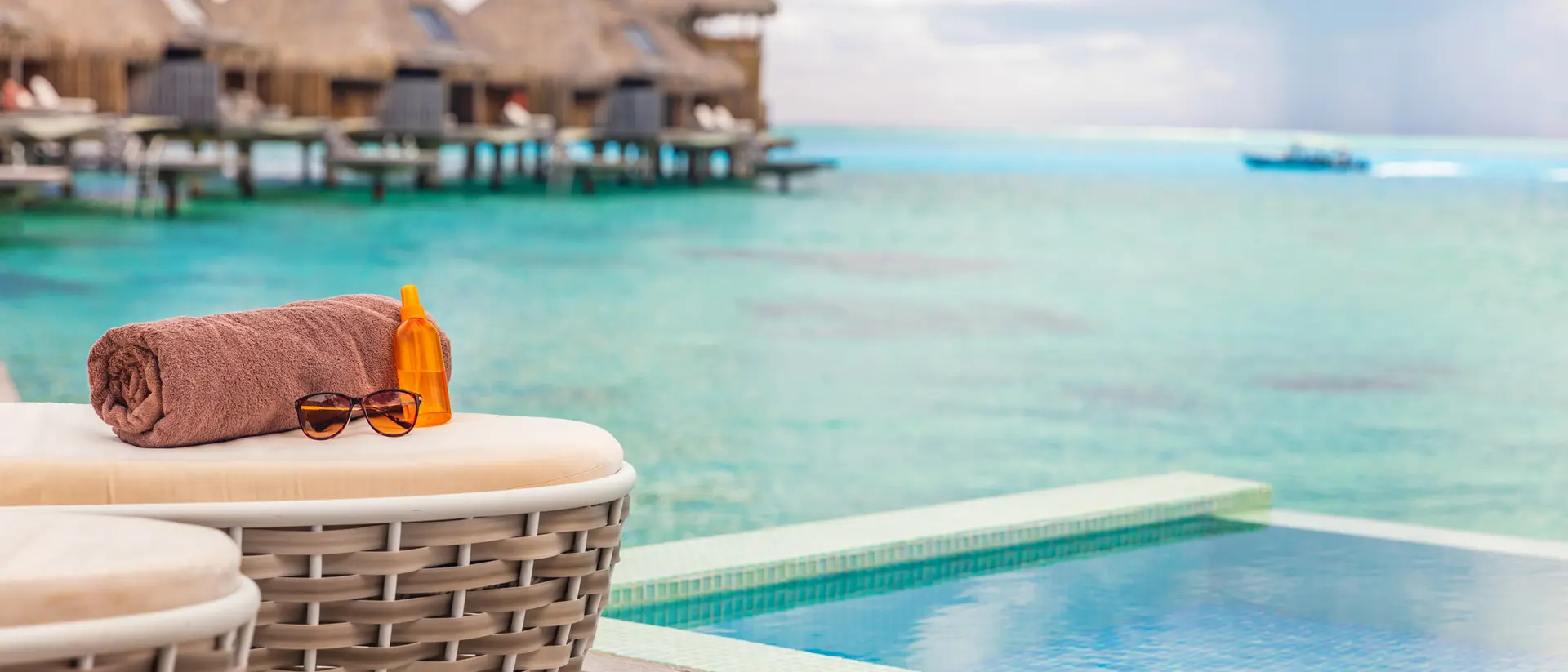 A poolside view featuring a rolled towel and sunglasses beside a turquoise sea and overwater bungalows in the background.