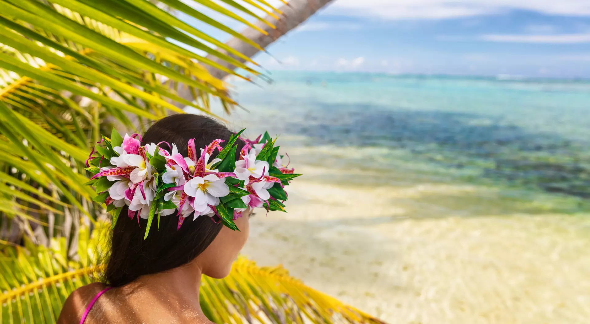 A woman with a flower crown gazes at a serene tropical beach with clear blue waters and palm leaves.