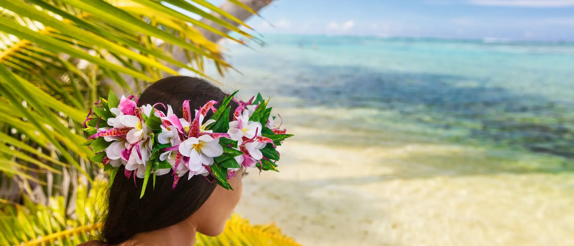 A woman with a flower crown gazes at a serene tropical beach with clear blue waters and palm leaves.