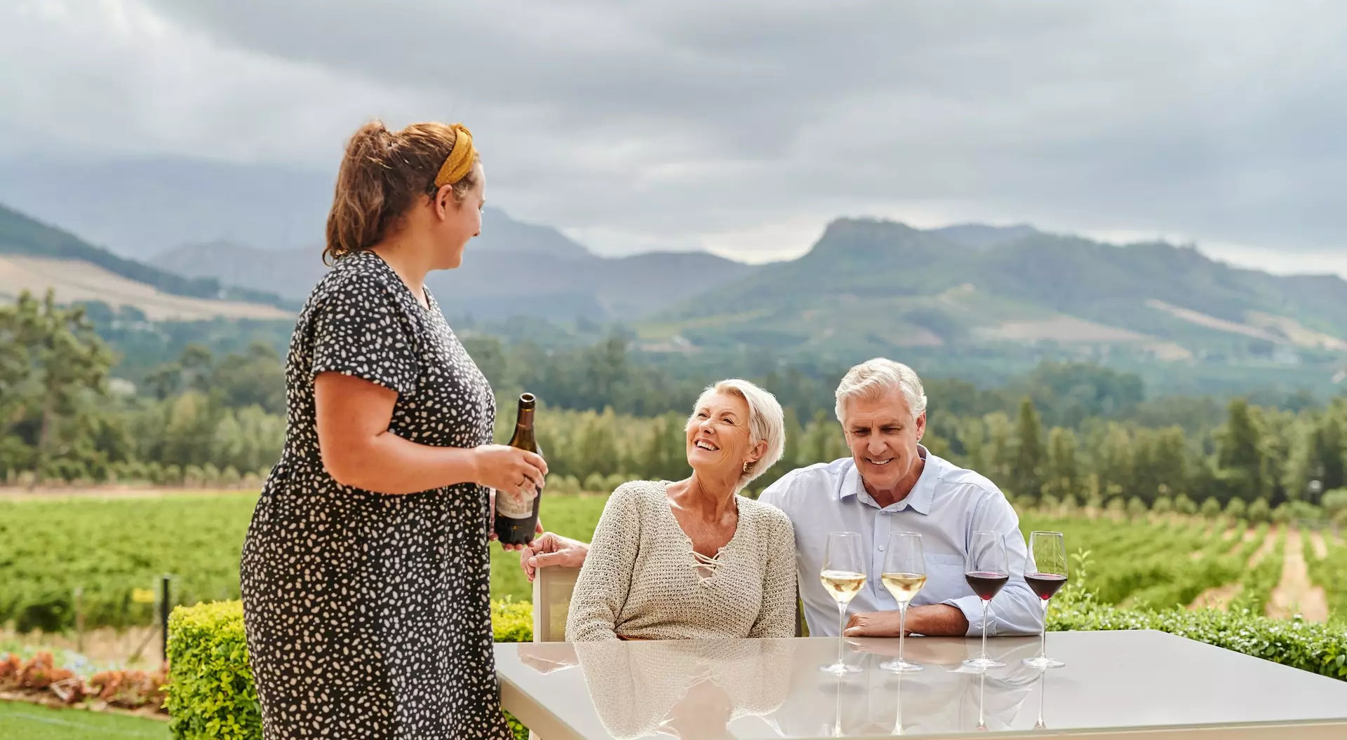 Three people enjoy a conversation over drinks at a vineyard table, with lush green hills in the background.