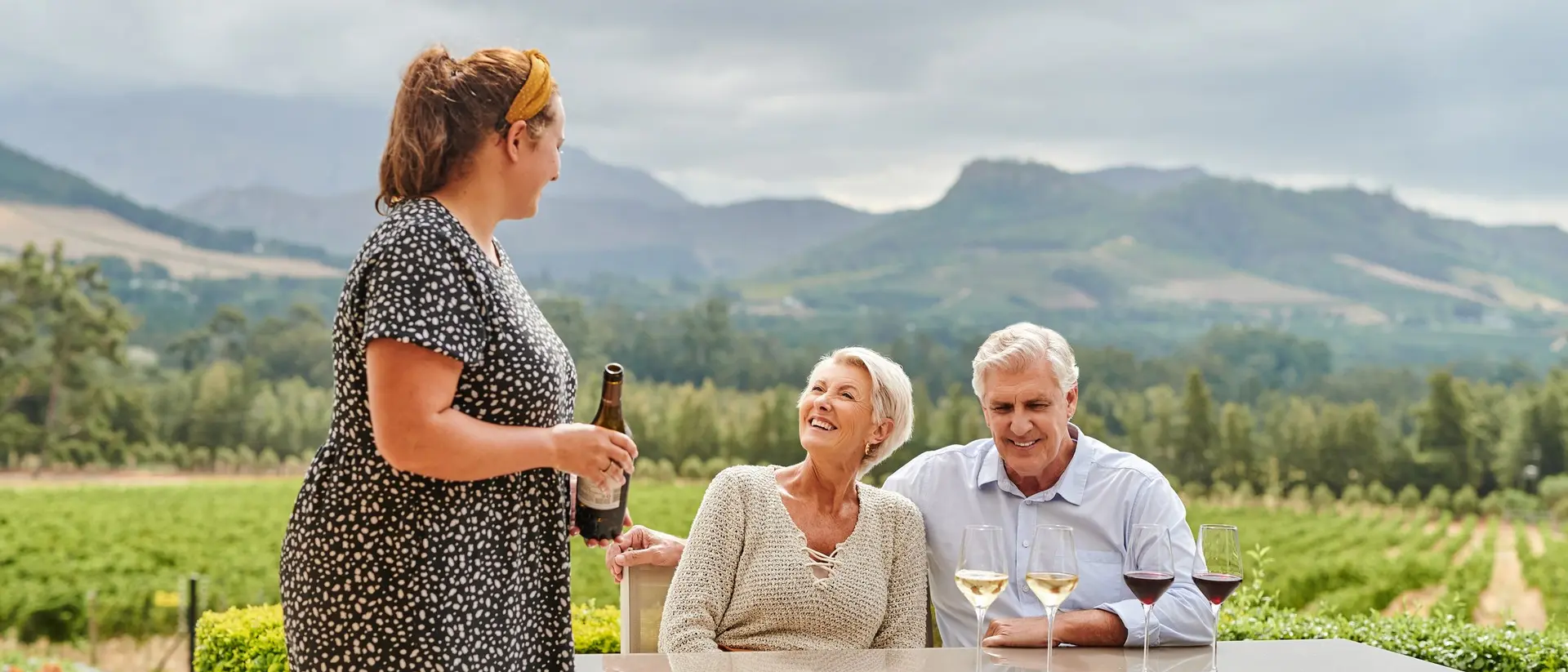 Three people enjoy a conversation over drinks at a vineyard table, with lush green hills in the background.