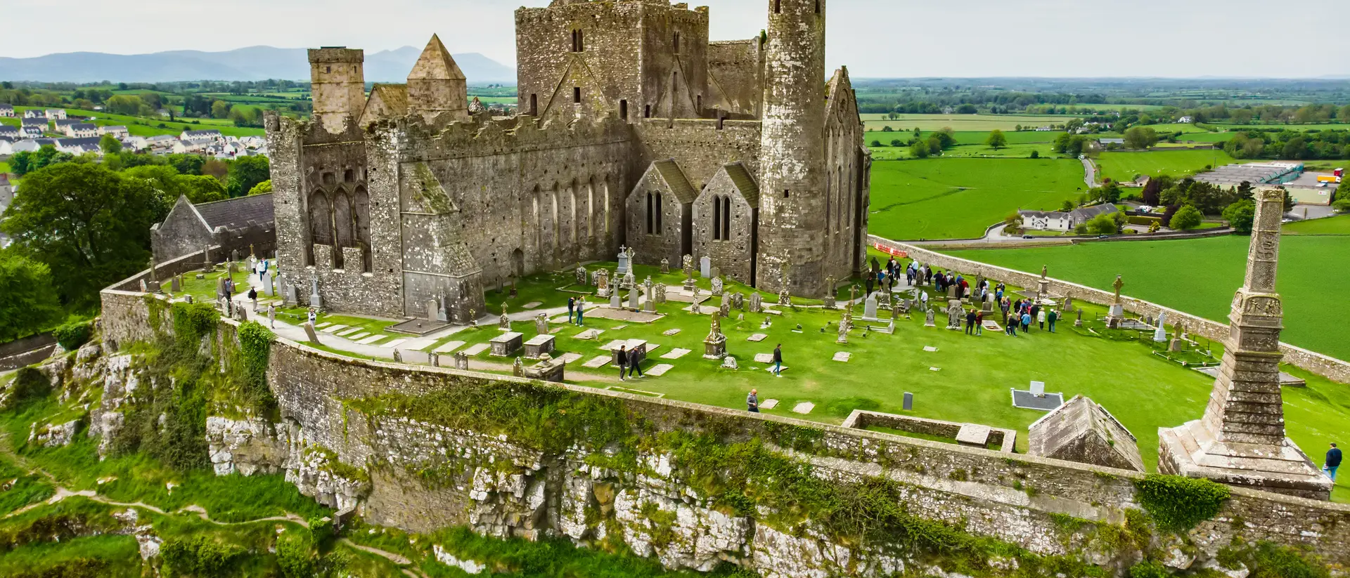 Aerial view of a historic castle surrounded by green fields and gravestones on a hilltop.