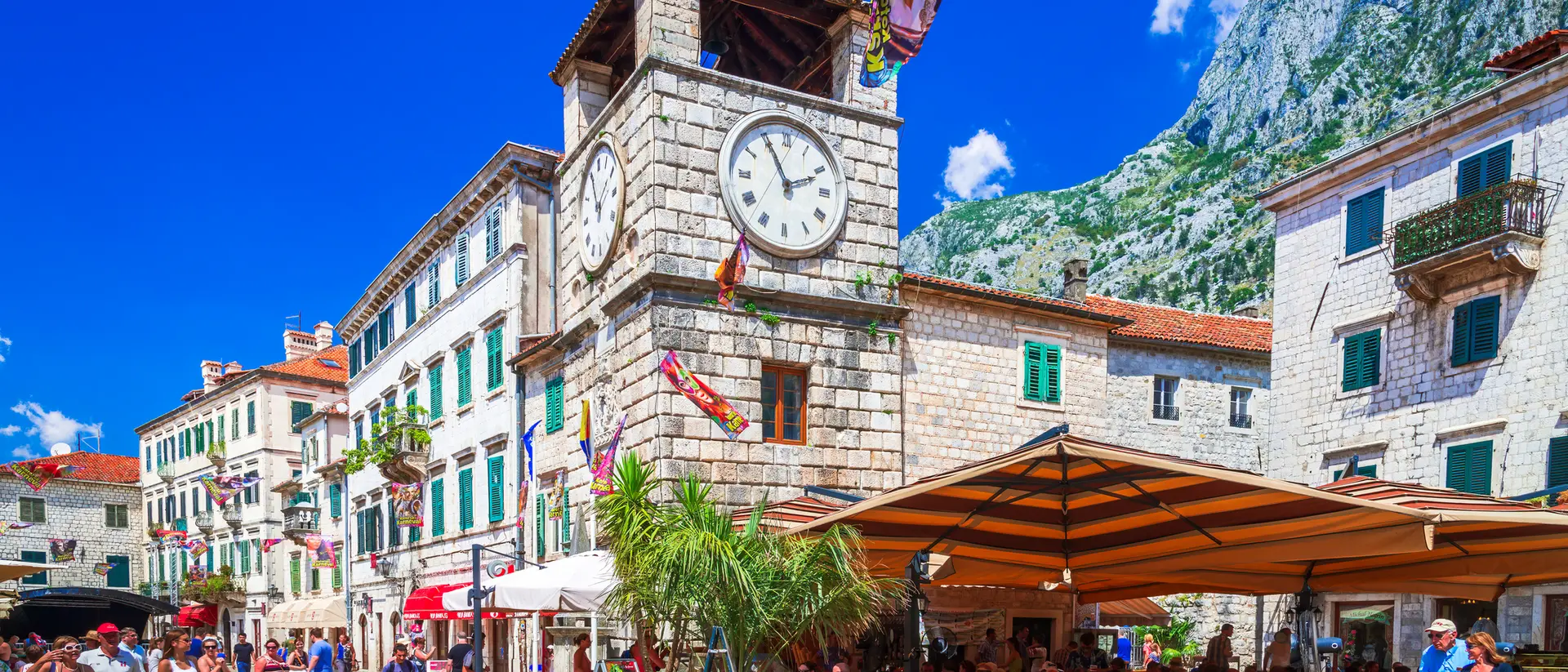 A bustling square in Kotor with a clock tower, cafes, and mountains under a clear blue sky.