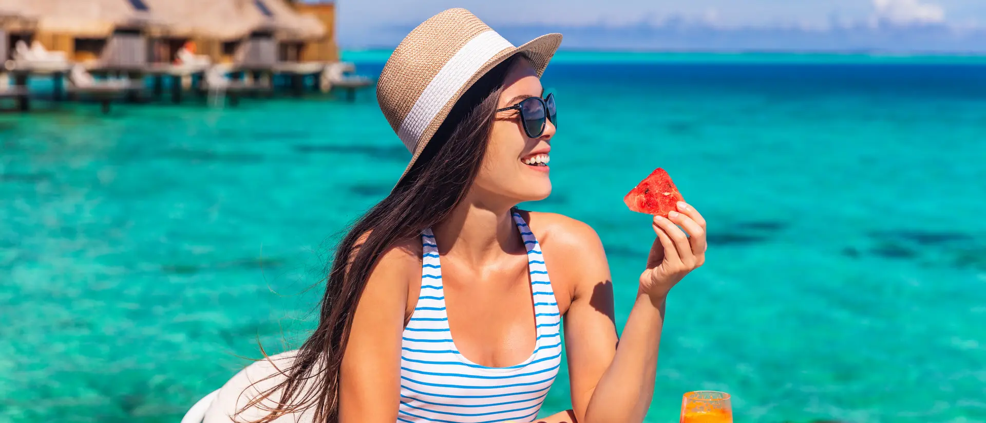 A woman in a striped swimsuit enjoys fresh fruit and a drink by the turquoise ocean, with overwater bungalows in the background.