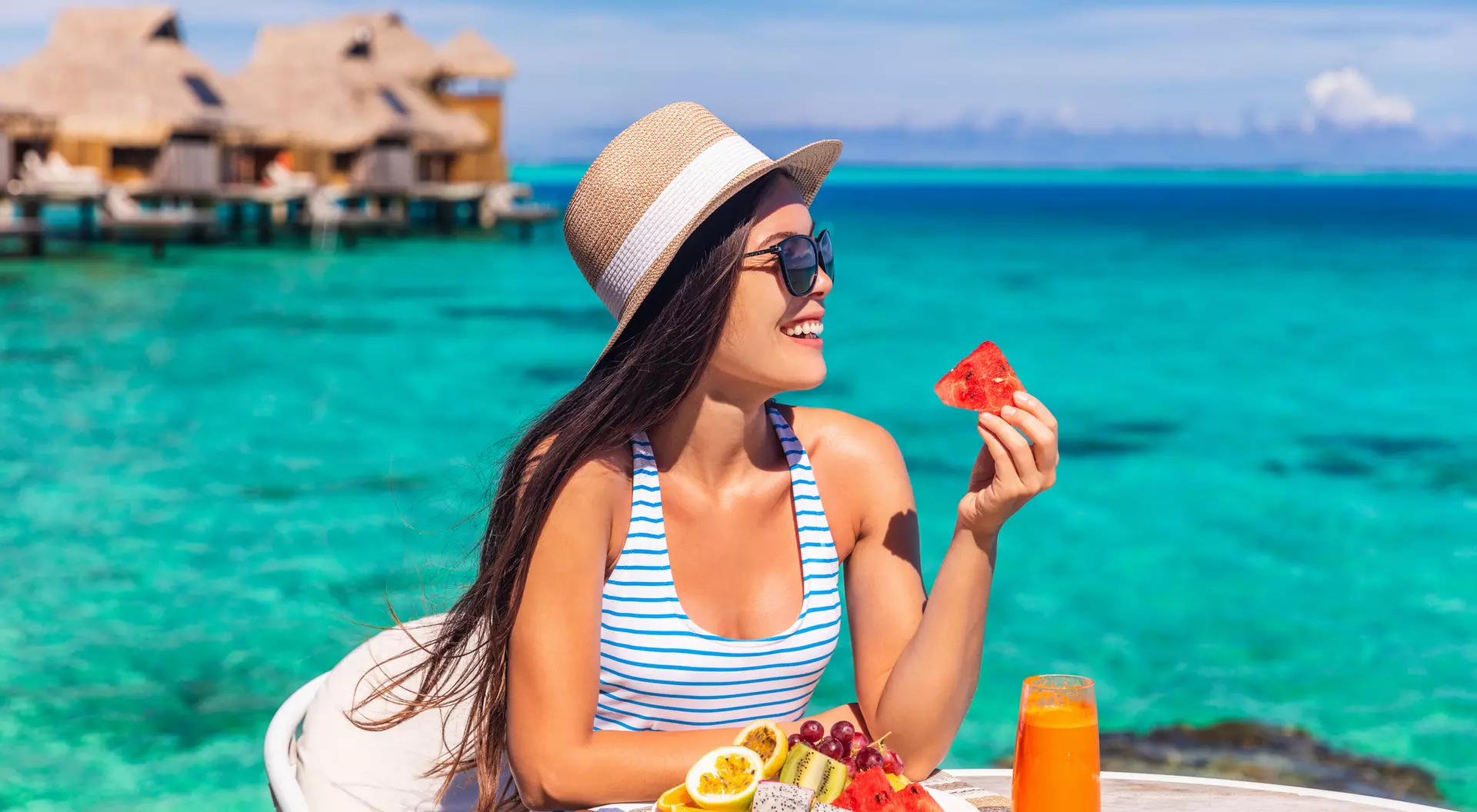 A woman in a striped swimsuit enjoys fresh fruit and a drink by the turquoise ocean, with overwater bungalows in the background.